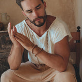 A man with a beard sits on a chair, dressed in a White Raw Edge Thick Cotton Top Tee by AYA Sacred Wear and cream-colored pants. His focused expression is accentuated by his hands clasped together, while a wooden necklace adorns his neck against the rustic backdrop of a wooden cabinet and a clay jug.