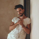 A man with a beard leans against a wall, wearing an AYA Sacred Wear White Raw Edge Thick Cotton Top Tee for Men paired with beige pants. He accessorizes with a long beaded necklace and bracelets, posing with hands together. The background is a textured, neutral-colored wall.