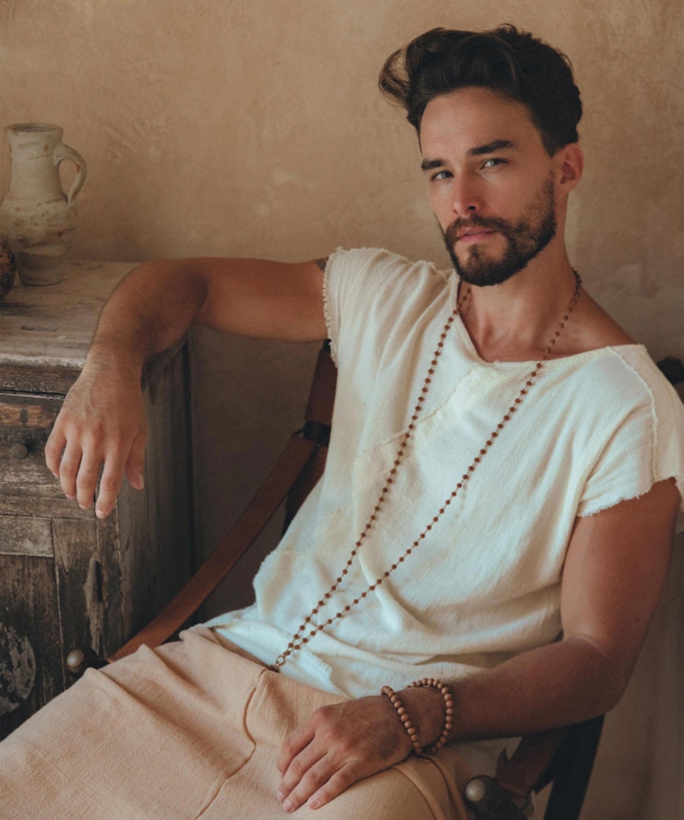A bearded man with dark hair is sitting on a chair, wearing an AYA Sacred Wear White Raw Edge Thick Cotton Top Tee for Men and beige pants. He has a wooden bead necklace and bracelet, set against the backdrop of a rustic wall and a wooden chest adorned with pottery.