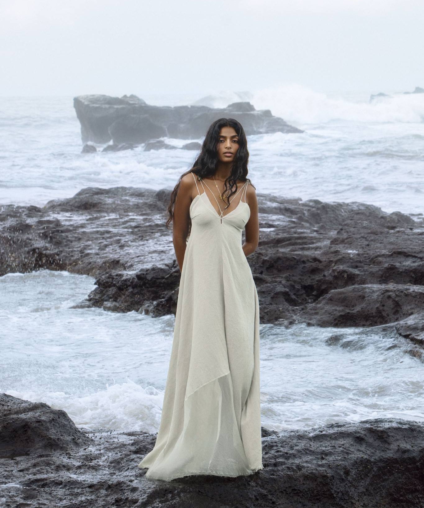A woman in an AYA Sacred Wear Powder Blue Padma Dress stands on rugged ocean rocks with crashing waves and an overcast sky. Her long black hair cascades down her shoulders as she gazes ahead.