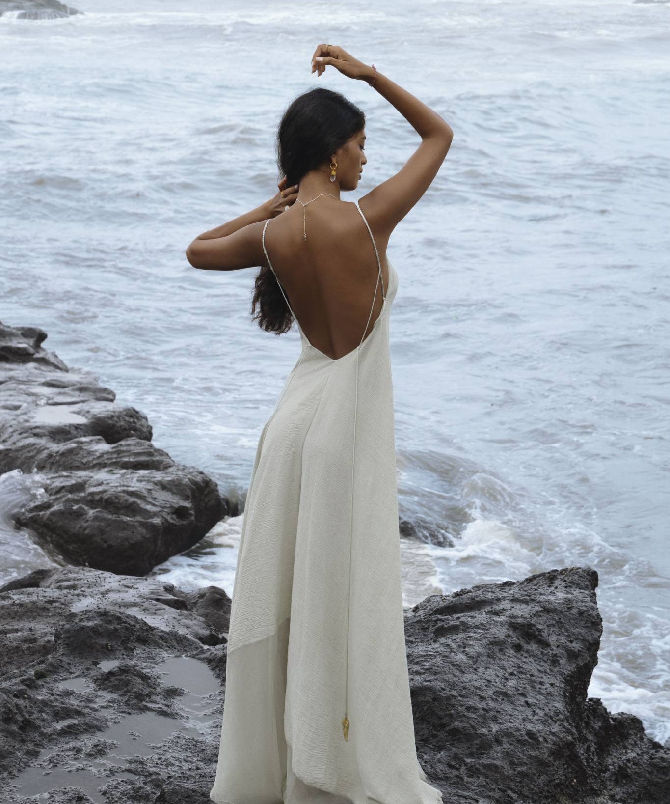 A woman wearing the Powder Blue Padma Dress by AYA Sacred Wear stands on rocky terrain by the ocean. Her long hair cascades down her back, and she raises one hand to touch her head, creating an elegant scene with waves in the background.