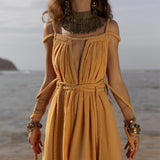 A woman stands on a beach in a flowing AYA Sacred Wear Yellow Gold Greek Goddess Prom Boho Dress featuring intricate rope details. She accessorizes with a large, ornate necklace and multiple bracelets against the ocean and distant hill, creating a stunning backdrop for this boho wedding moment.