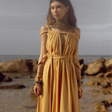 On a rocky beach under a cloudy sky, a woman wears the Yellow Gold Greek Goddess Prom Boho Dress by AYA Sacred Wear. The long, flowing dress, rich with intricate braided details, complements her ornate jewelry, as the ocean and rocks form the scenic backdrop.