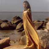A woman in the Yellow Gold Greek Goddess Prom Boho Dress by AYA Sacred Wear stands barefoot on rocks by the sea. Her long dark hair flows as she gazes into the distance, with an overcast sky above and a calm ocean forming a serene backdrop. Perfect for special occasions.