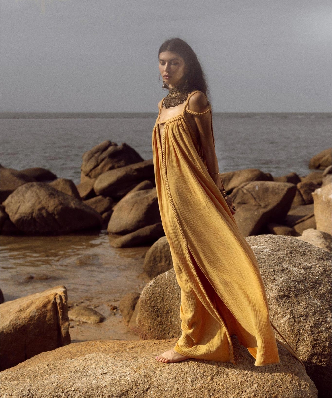 A woman in the Yellow Gold Greek Goddess Prom Boho Dress by AYA Sacred Wear stands barefoot on rocks by the sea. Her long dark hair flows as she gazes into the distance, with an overcast sky above and a calm ocean forming a serene backdrop. Perfect for special occasions.