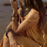 A woman in an AYA Sacred Wear Yellow Gold Greek Goddess Prom Boho Dress sits by the beach, adorned with intricate jewelry including a necklace, hoop earrings, and bracelets. Her long brown hair cascades down her back as she gazes upwards.