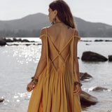 A woman in a flowing backless "Yellow Gold Greek Goddess Prom Boho Dress" by AYA Sacred Wear stands on rocks by the water. Facing away, mountains form her backdrop as large earrings and bracelets glint in the sun, reflecting off the shimmering surface and embodying boho wedding dress elegance.