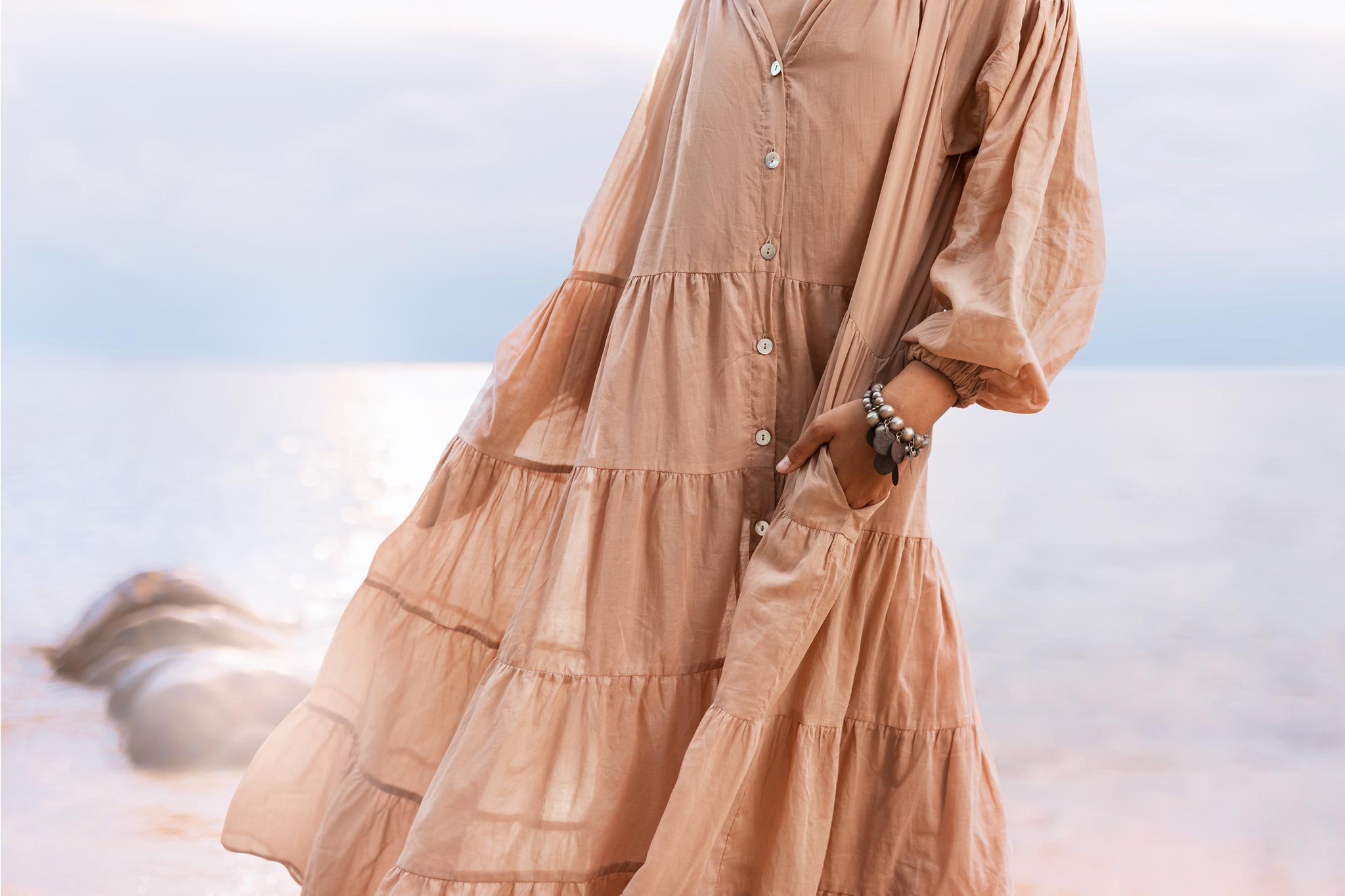 A person in a flowing dusty pink Boho Dress by AYA Sacred Wear stands by the water's edge, hand in pocket. This cotton maxi dress features buttons and layered tiers, against a serene beach backdrop with soft sunlight dancing on the water.
