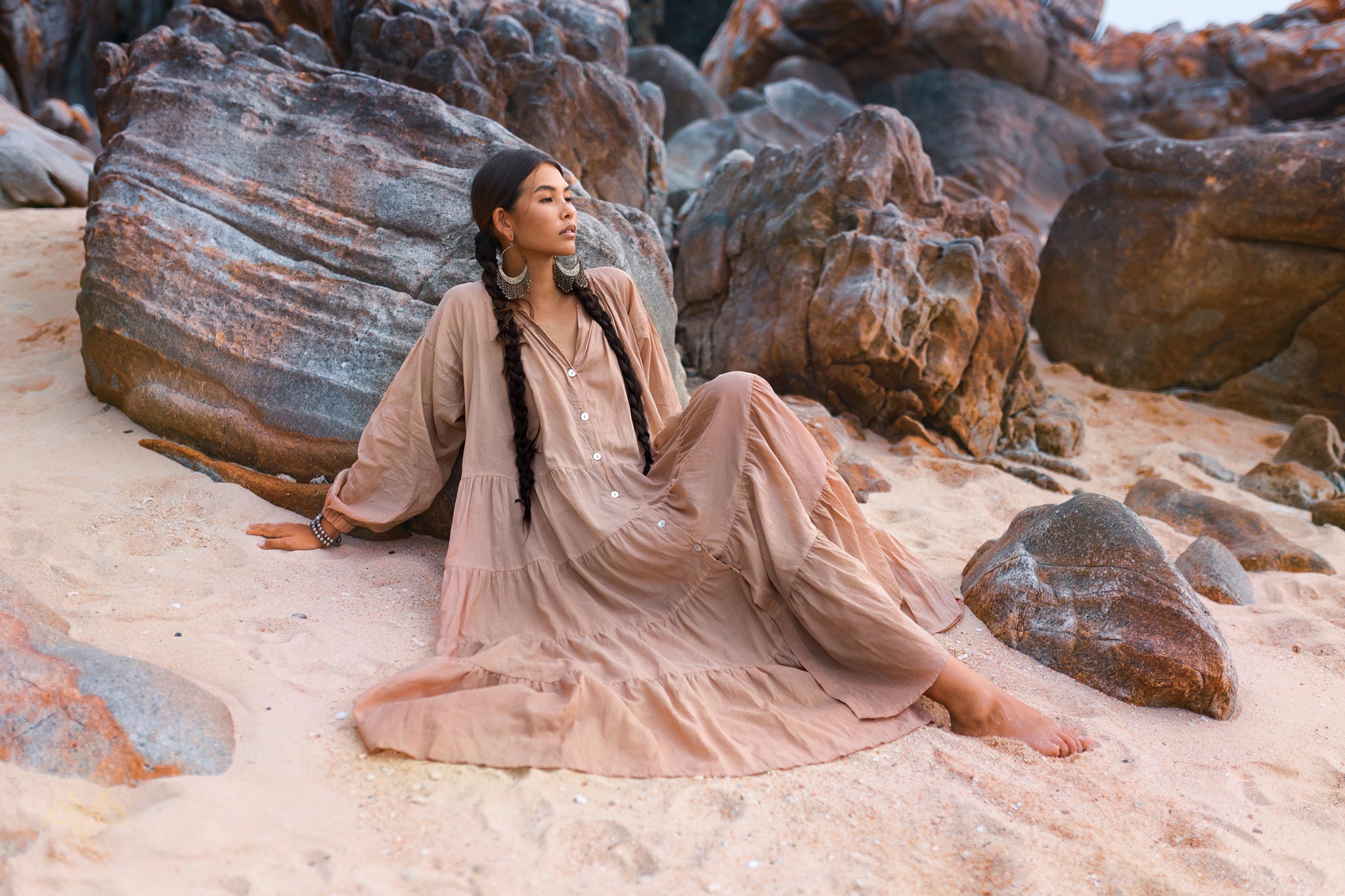 A woman in a dusty pink Boho maxi dress by AYA Sacred Wear sits on sandy ground, leaning against large rocks on the beach. Her long braided hair cascades down her back as she gazes thoughtfully into the distance, creating a serene and natural scene.