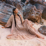 A woman in a dusty pink Boho maxi dress by AYA Sacred Wear sits on sandy ground, leaning against large rocks on the beach. Her long braided hair cascades down her back as she gazes thoughtfully into the distance, creating a serene and natural scene.