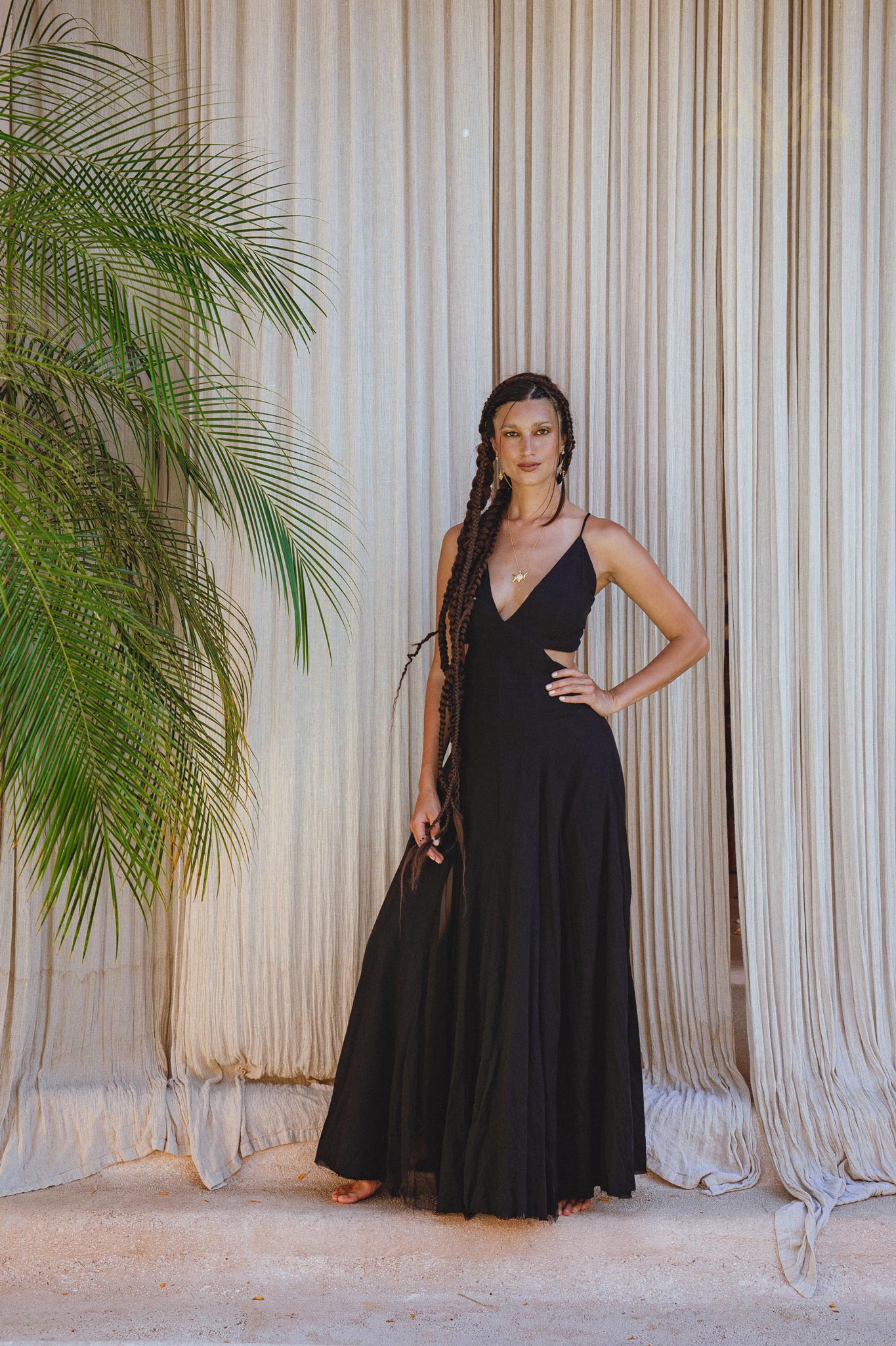 A woman poses barefoot indoors in front of beige curtains and a leafy plant, wearing the AYA Sacred Wear Black Oshun Dress—a long, black A-line formal bridal dress crafted from light cotton and silk.
