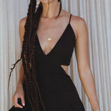 A woman with long, braided hair wears the Black Oshun Dress by AYA Sacred Wear—a sleeveless A-line formal dress in light cotton and silk with a deep V-neck—paired with a gold pendant necklace, standing before light curtains.