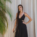 A woman with long braided hair smiles softly as she poses in front of beige curtains and a leafy plant, wearing the Black Oshun Dress by AYA Sacred Wear—a sleeveless, formal A-line dress in light cotton and silk with cut-out detailing.