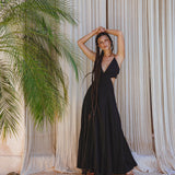 A woman with long braids smiles barefoot in front of beige curtains and a green plant, wearing the Black Oshun Dress—a light cotton and silk A-line formal bridal dress by AYA Sacred Wear, her arms raised above her head.