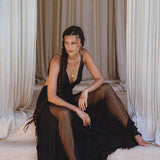 A woman with long braids sits barefoot before cream curtains, wearing the Black Oshun Dress by AYA Sacred Wear—a sleeveless black A-line dress in light cotton and silk—with a gold necklace, gazing thoughtfully to the side.