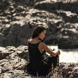A woman in an AYA Sacred Wear Black Cotton Bohemian Dress sits on rocky terrain, facing away but glancing over her shoulder as sunlight illuminates her face and the textured rocks around her.