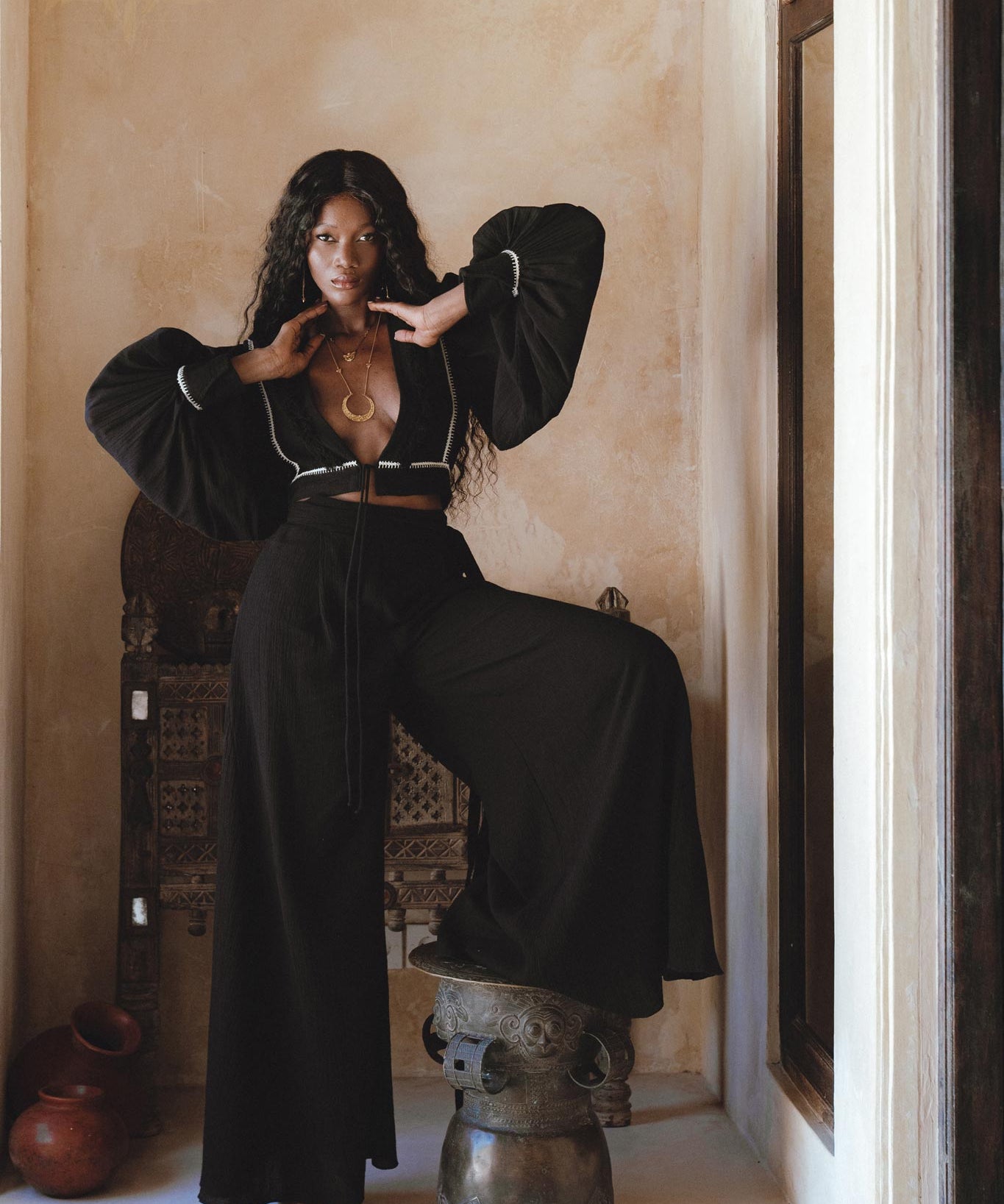 A model showcases black bohemian craft trousers from Sacred Wear, styled with a matching black cropped top. The outfit features voluminous sleeves and a relaxed fit, complemented by layered gold jewelry. The model poses confidently with one leg elevated on a decorative stool, against a warm-toned wall and elegant background elements, highlighting a casual boho aesthetic. Perfect for boho casual occasions.