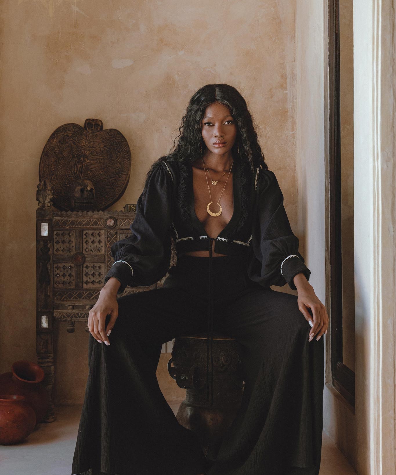 A model sitting on a decorative stool, wearing black boho pants and a matching cropped top. The outfit features flowing fabric and a relaxed fit, complemented by layered gold necklaces. The backdrop includes earthy tones and rustic decor, enhancing the bohemian aesthetic. Keywords: black boho pants, boho casual trousers, bohemian craft trousers by AYA Sacred Wear.
