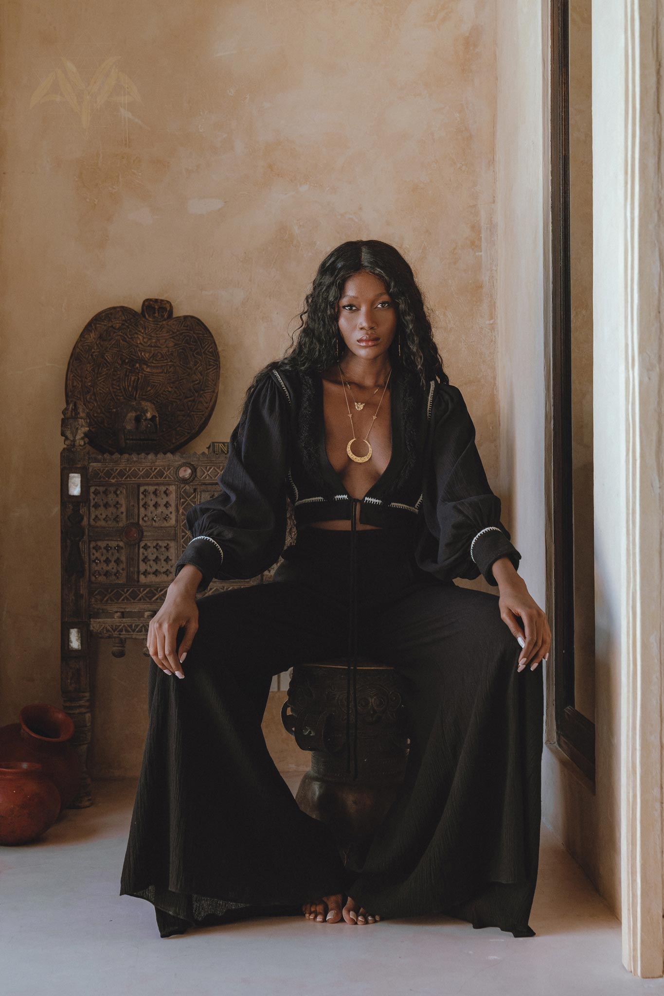 A model sitting on a decorative stool, wearing black boho pants and a matching cropped top. The outfit features flowing fabric and a relaxed fit, complemented by layered gold necklaces. The backdrop includes earthy tones and rustic decor, enhancing the bohemian aesthetic. Keywords: black boho pants, boho casual trousers, bohemian craft trousers by AYA Sacred Wear.