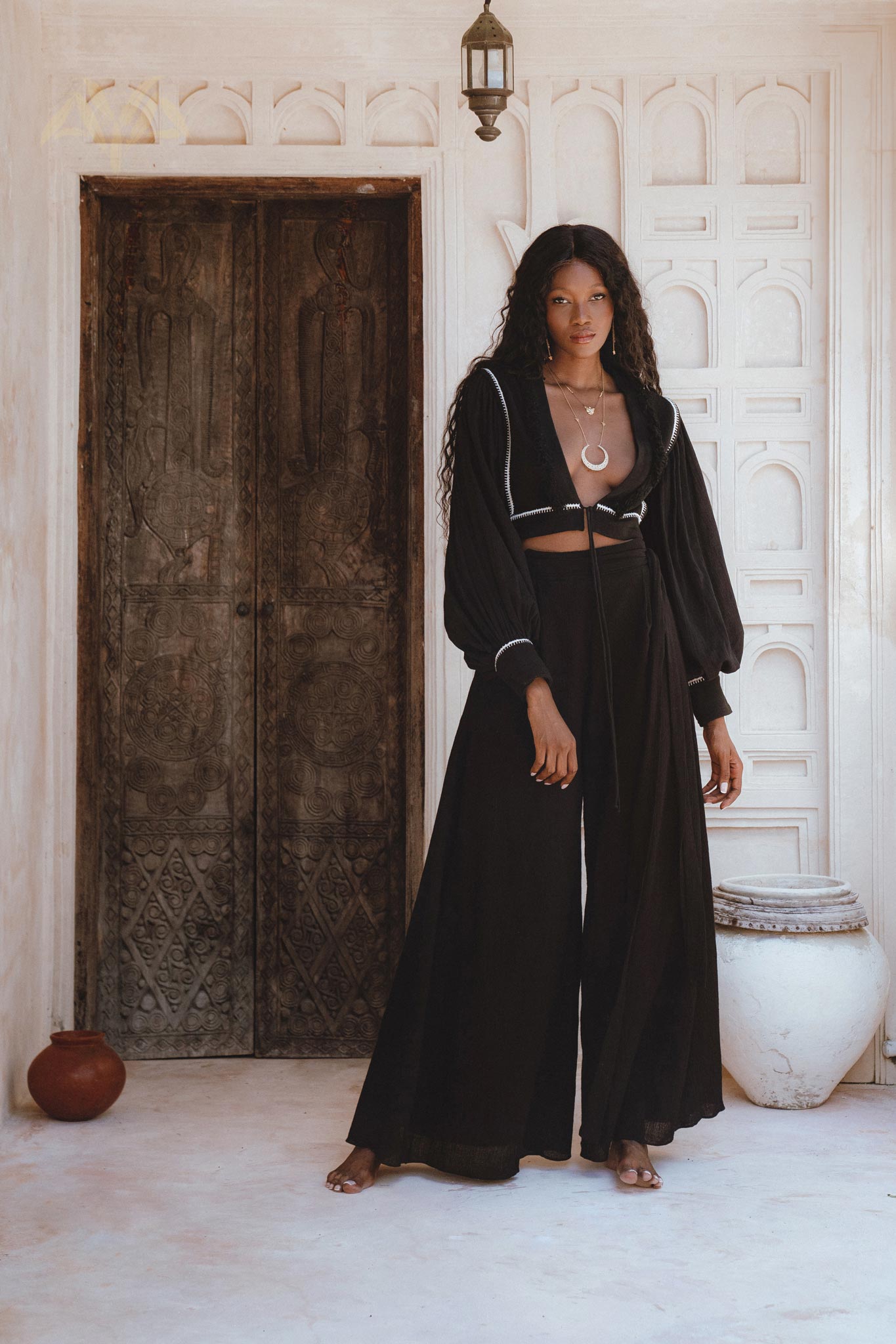 A model showcasing black boho pants, styled with a matching top, set against a rustic backdrop featuring ornate wooden doors. The outfit embodies a bohemian aesthetic, perfect for casual wear.
