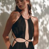 A woman showcases the Black Prom Handmade Dress from AYA Sacred Wear, a boho dress with cut-out details crafted from organic fabric. She stands against a wall playfully illuminated by sunlight and shadow, gazing downward, with her long wavy hair cascading over her shoulder.