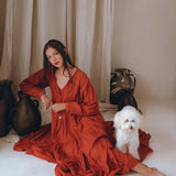 A woman wearing an AYA Sacred Wear Wine Red Boho Prom Dress gracefully sits on the floor against a beige curtain backdrop. Next to her is a small, fluffy white dog, with dark decorative vases arranged nearby.
