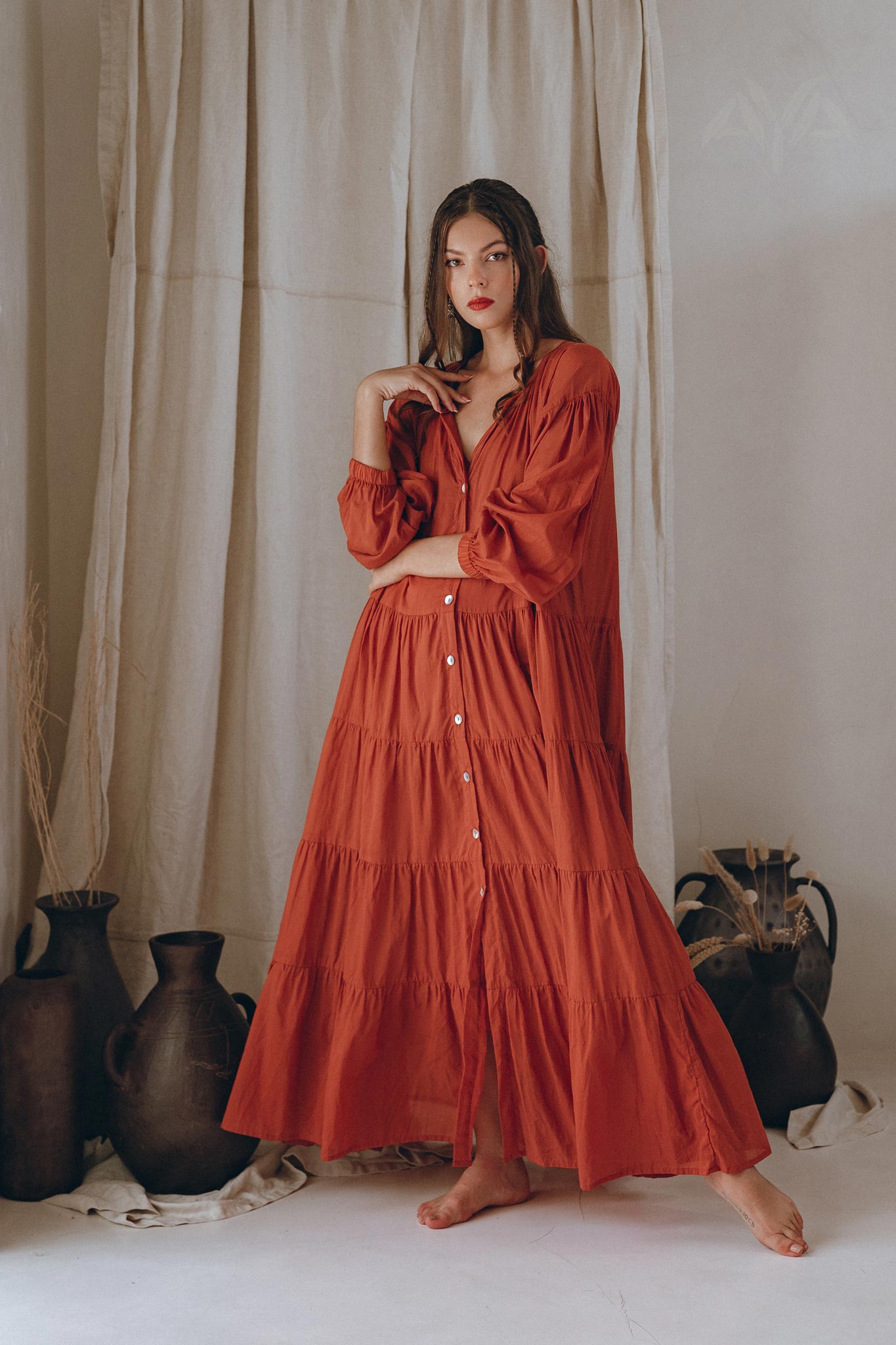 A woman wearing a Boho Dress for Women by AYA Sacred Wear, specifically the Wine Red Boho Prom Dress, stands barefoot in front of beige curtains. She gracefully holds part of her cotton Light Summer Dress with one hand, as ceramic pots rest on the floor beside her. Her long hair flows freely.