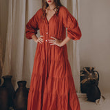 A woman stands confidently in a flowing Wine Red Boho Prom Dress with long sleeves, from AYA Sacred Wear. Her hands are on her hips, exuding poise. The background features neutral-toned curtains and decorative vases, enhancing her stylish presence in this Boho Dress for Women.