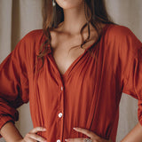 A woman with long brown hair and red lipstick stands confidently with her hands on her hips. She is wearing an AYA Sacred Wear Boho Dress for Women in a flowing wine red color, adorned with buttons and crafted from elegant cotton fabric. Complementing her look are silver earrings, while the background features softly draped fabric.