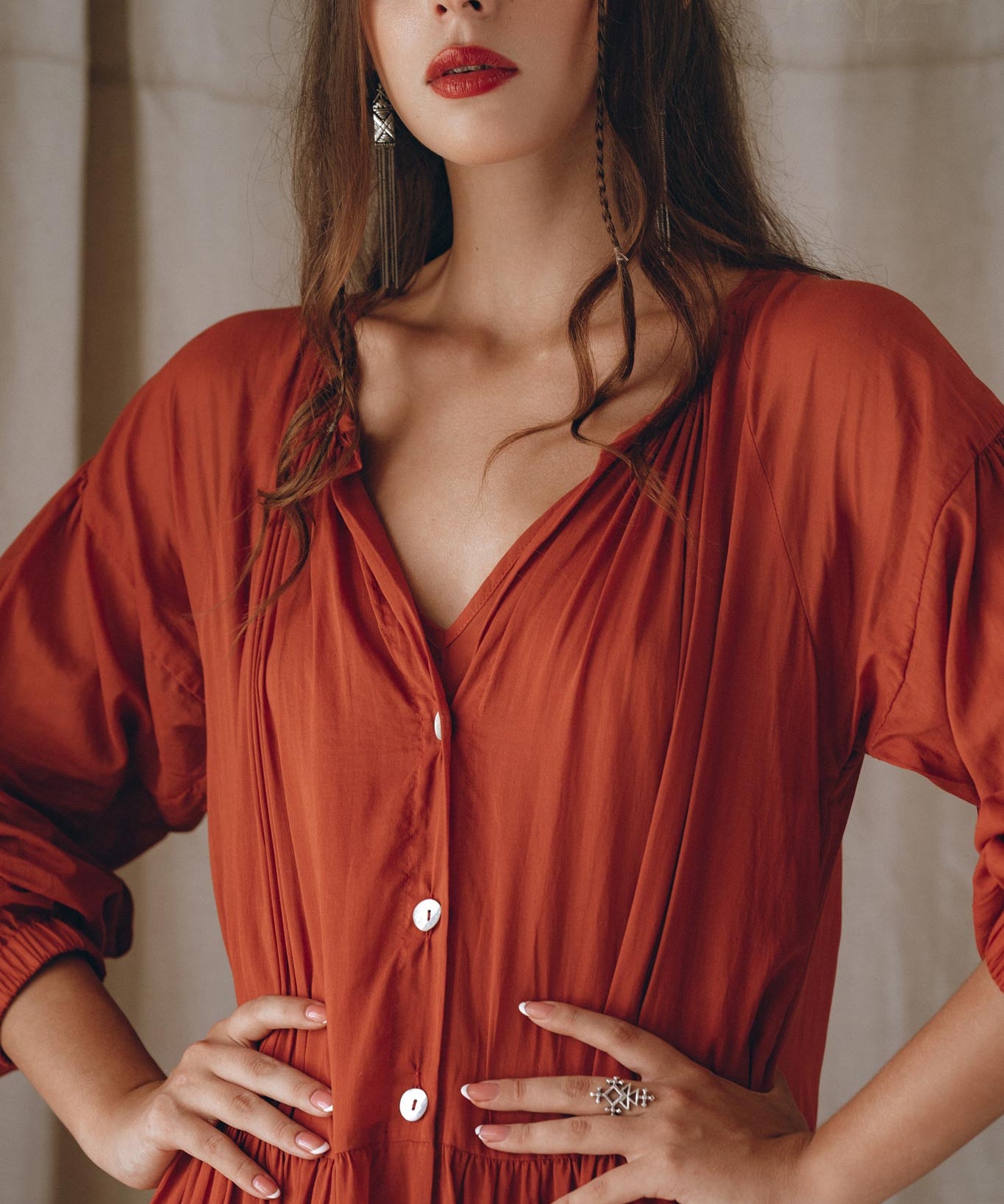 A woman with long brown hair and red lipstick stands confidently with her hands on her hips. She is wearing an AYA Sacred Wear Boho Dress for Women in a flowing wine red color, adorned with buttons and crafted from elegant cotton fabric. Complementing her look are silver earrings, while the background features softly draped fabric.