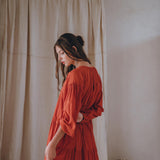 A woman wearing the AYA Sacred Wear Wine Red Boho Prom Dress stands sideways in a softly lit room. Her hair is tied back as she gazes downward, surrounded by a neutral-toned curtain and pottery with dried plants.