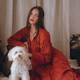 A woman in a beautiful Wine Red Boho Prom Dress from AYA Sacred Wear sits on the floor next to a fluffy white dog. The background features neutral-colored curtains and decorative pottery, creating a warm, elegant atmosphere.