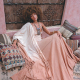 A woman with curly hair relaxes on patterned cushions in a flowy Powder Pink Boho Poncho by AYA Sacred Wear, crafted sustainably with ombre botanical dyes. She's enveloped by rustic wooden doors and textured textiles, creating a bohemian ambiance.