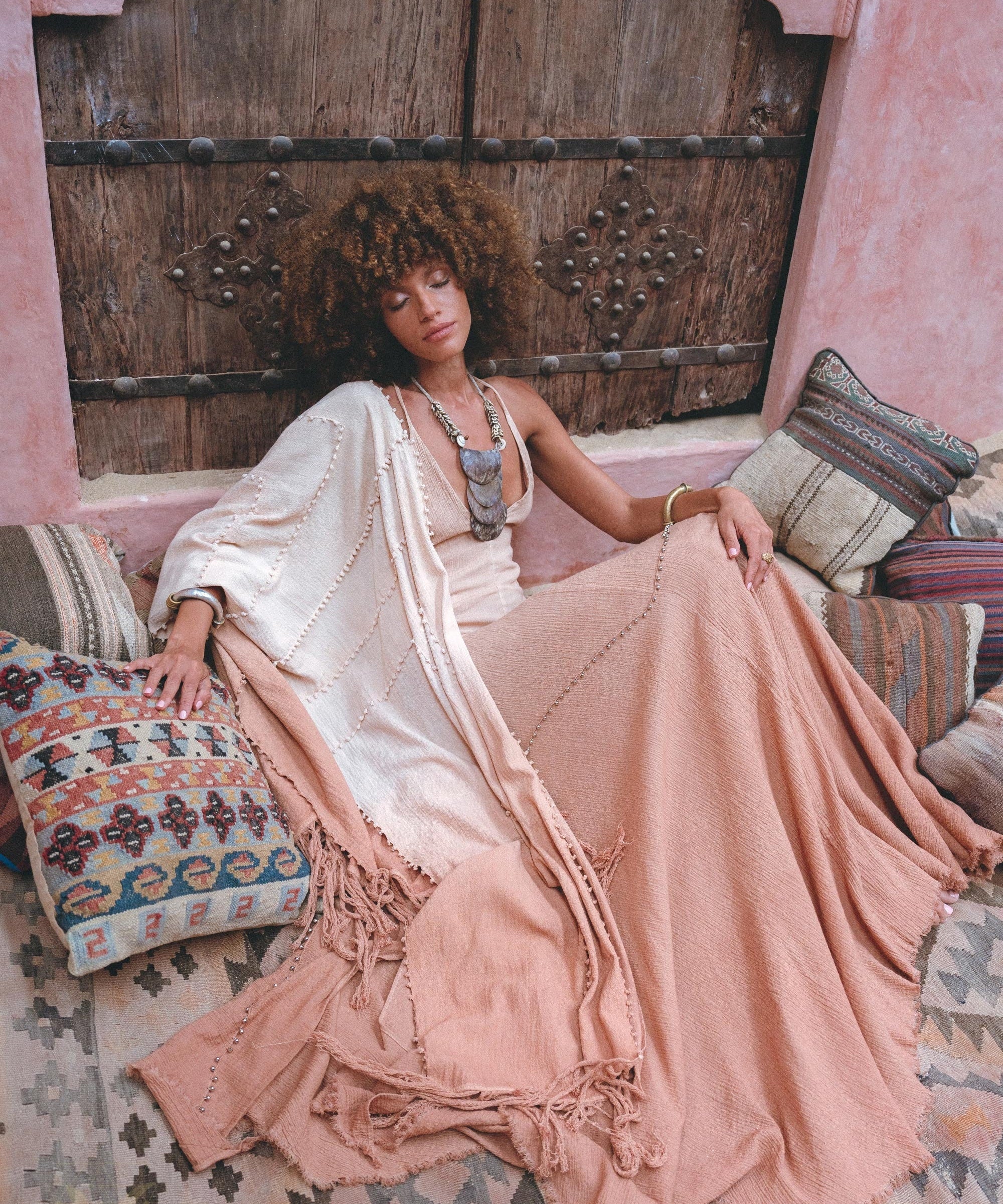 A woman with curly hair relaxes on patterned cushions in a flowy Powder Pink Boho Poncho by AYA Sacred Wear, crafted sustainably with ombre botanical dyes. She's enveloped by rustic wooden doors and textured textiles, creating a bohemian ambiance.