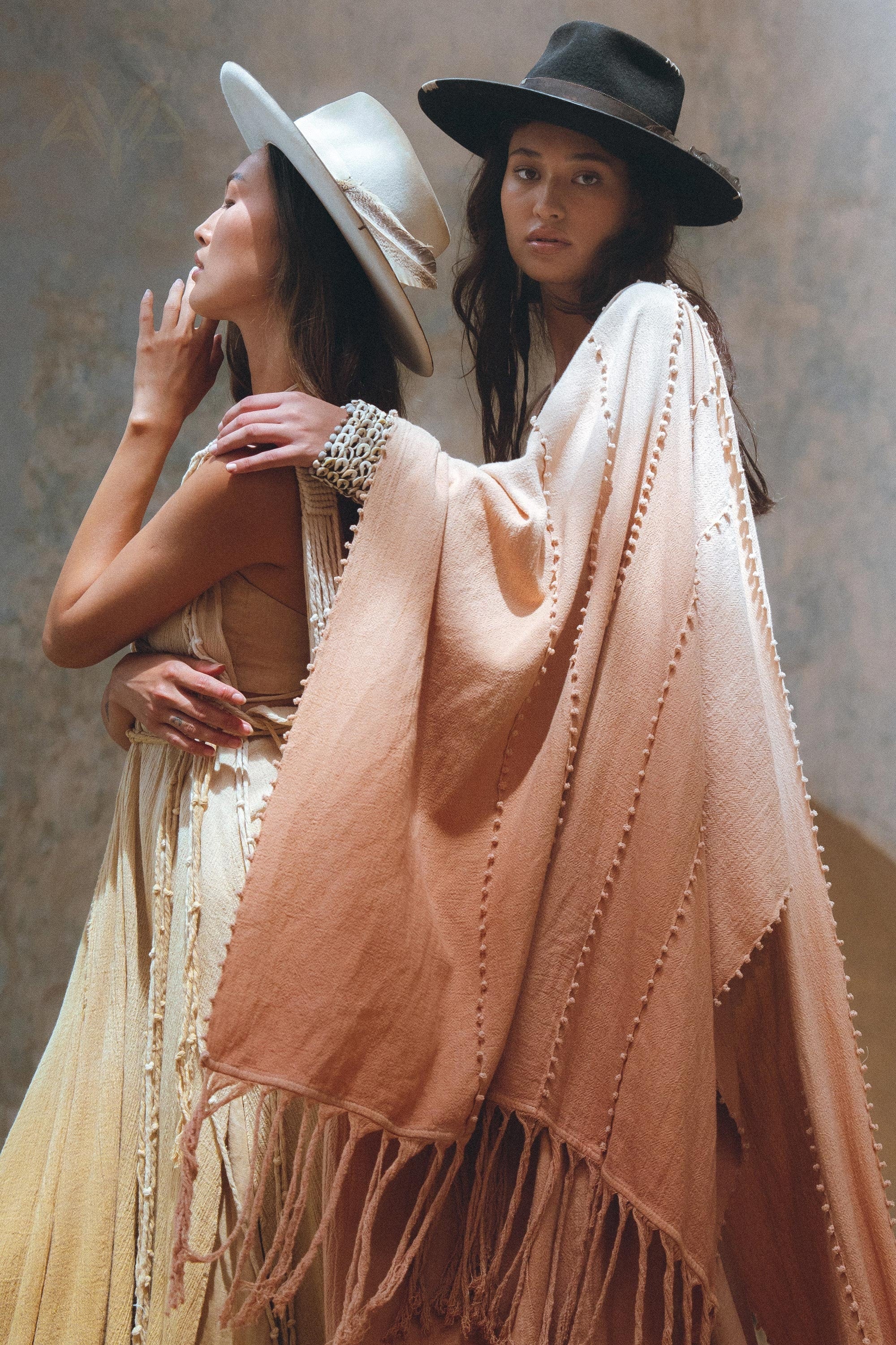 Two women in bohemian-style clothing and wide-brimmed hats. The woman on the left wears a flowing, sleeveless dress. On the right, a woman draped in AYA Sacred Wear's Powder Pink Boho Poncho rests her arm on her companion's shoulder.