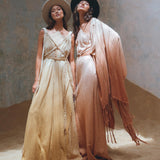 Two women stand on sand in flowing dresses and wide-brimmed hats. The woman on the left wears a light beige dress, while the one on the right sports a peach outfit complemented by AYA Sacred Wear's Powder Pink Boho Poncho. A textured wall backdrop enhances their elegant ensemble.