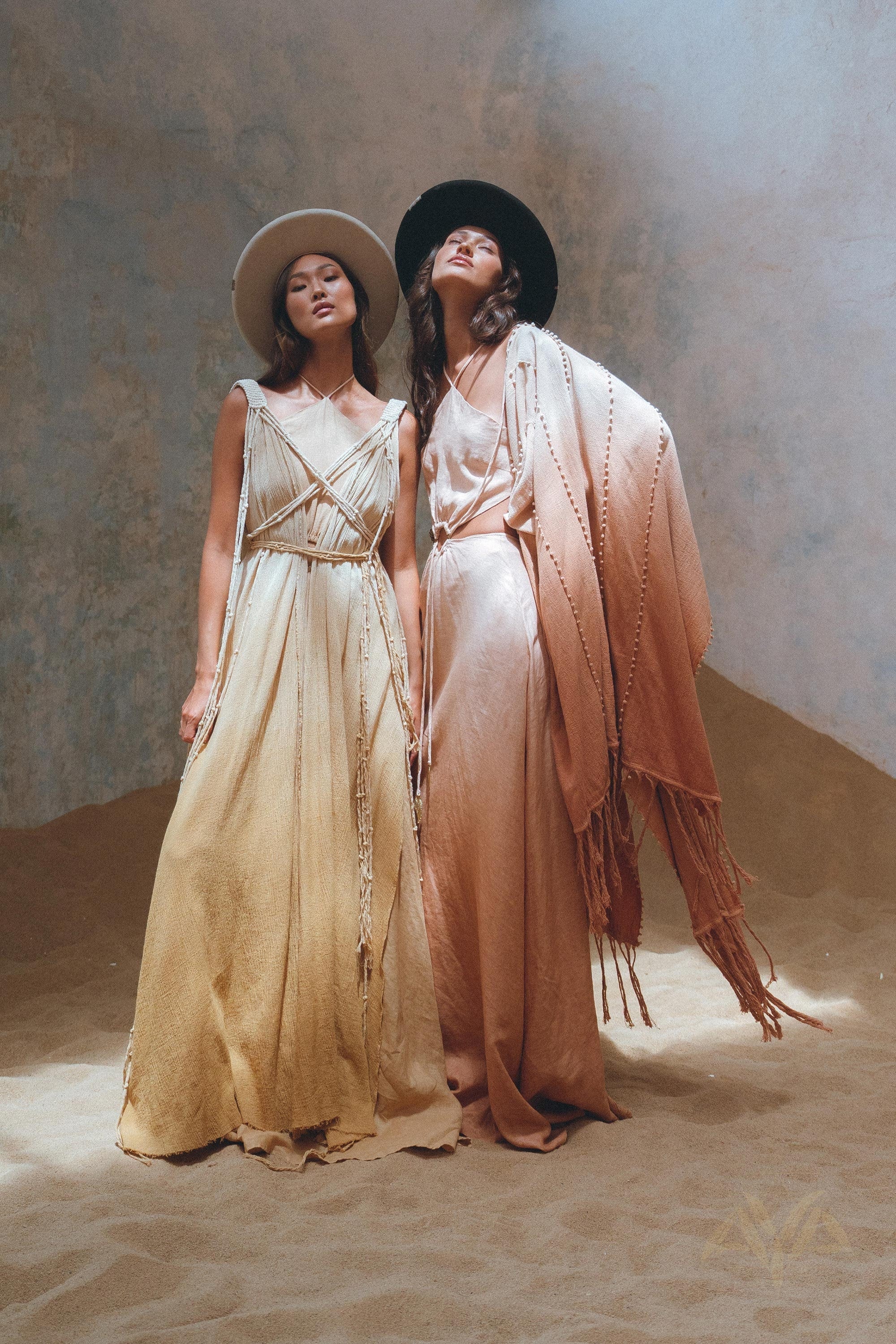 Two women stand on sand in flowing dresses and wide-brimmed hats. The woman on the left wears a light beige dress, while the one on the right sports a peach outfit complemented by AYA Sacred Wear's Powder Pink Boho Poncho. A textured wall backdrop enhances their elegant ensemble.