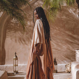 A person with long braided hair stands outside in a sunlit courtyard, wearing an AYA Sacred Wear Powder Pink Boho Poncho crafted from botanical dyeing techniques. They are surrounded by lush palm plants and decorative lanterns that cast soft shadows on the pale wall.