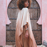 A person with curly hair stands before a carved wooden door in a pink, arched wall, wearing a Powder Pink Boho Poncho by AYA Sacred Wear, featuring cream and peach hues. They're surrounded by patterned cushions.