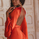 A woman poses gracefully in a wine red maxi dress with an open back, featuring long sleeves and a flowing silhouette. She is standing against a textured beige backdrop, showcasing the elegant design of the dress, perfect for a beach wedding. The look is completed with statement earrings and a stylish accessory on her wrist.