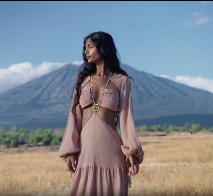 A woman with long dark hair stands in a golden field, wearing the AYA Sacred Wear Samudra Dress in Blush Rose—a backless boho maxi dress. She faces left, with distant mountains, trees, and a blue sky with scattered clouds behind her.