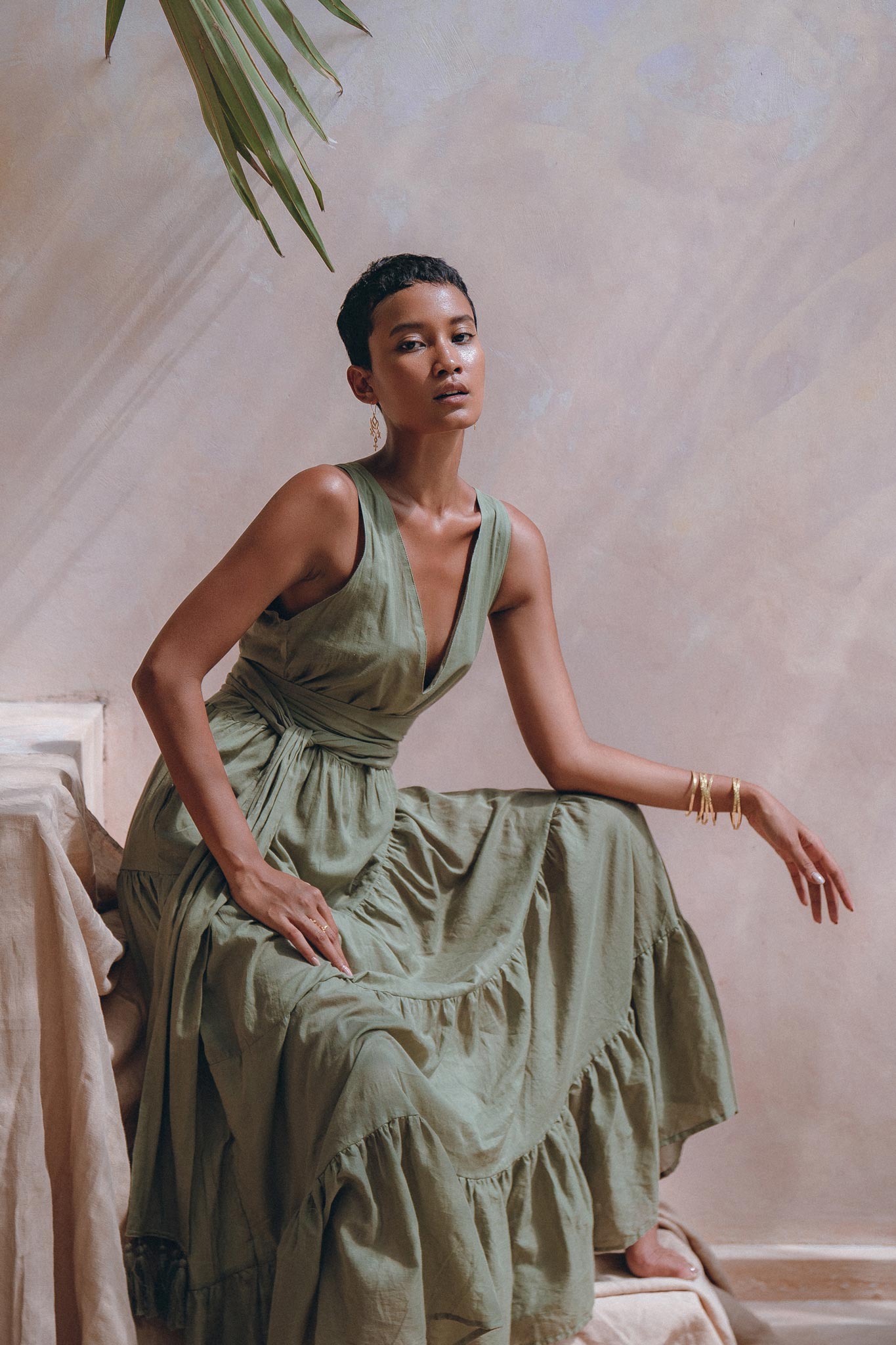 A woman in an AYA Sacred Wear Boho Bridesmaid Dress, a stylish Sage Green Adjustable Bohemian Dress, sits elegantly on a textured surface. She accessorizes with gold hoop earrings and bracelets, set against the backdrop of softly hued earth-toned walls and a green leaf overhead, creating a serene atmosphere.