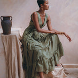A woman in a flowing Sage Green Adjustable Dress from AYA Sacred Wear sits gracefully on a draped surface, next to a decorative vase. Her posture is elegant, and the soft lighting adds a serene ambiance. A large green leaf hangs above, enhancing the earthy setting.