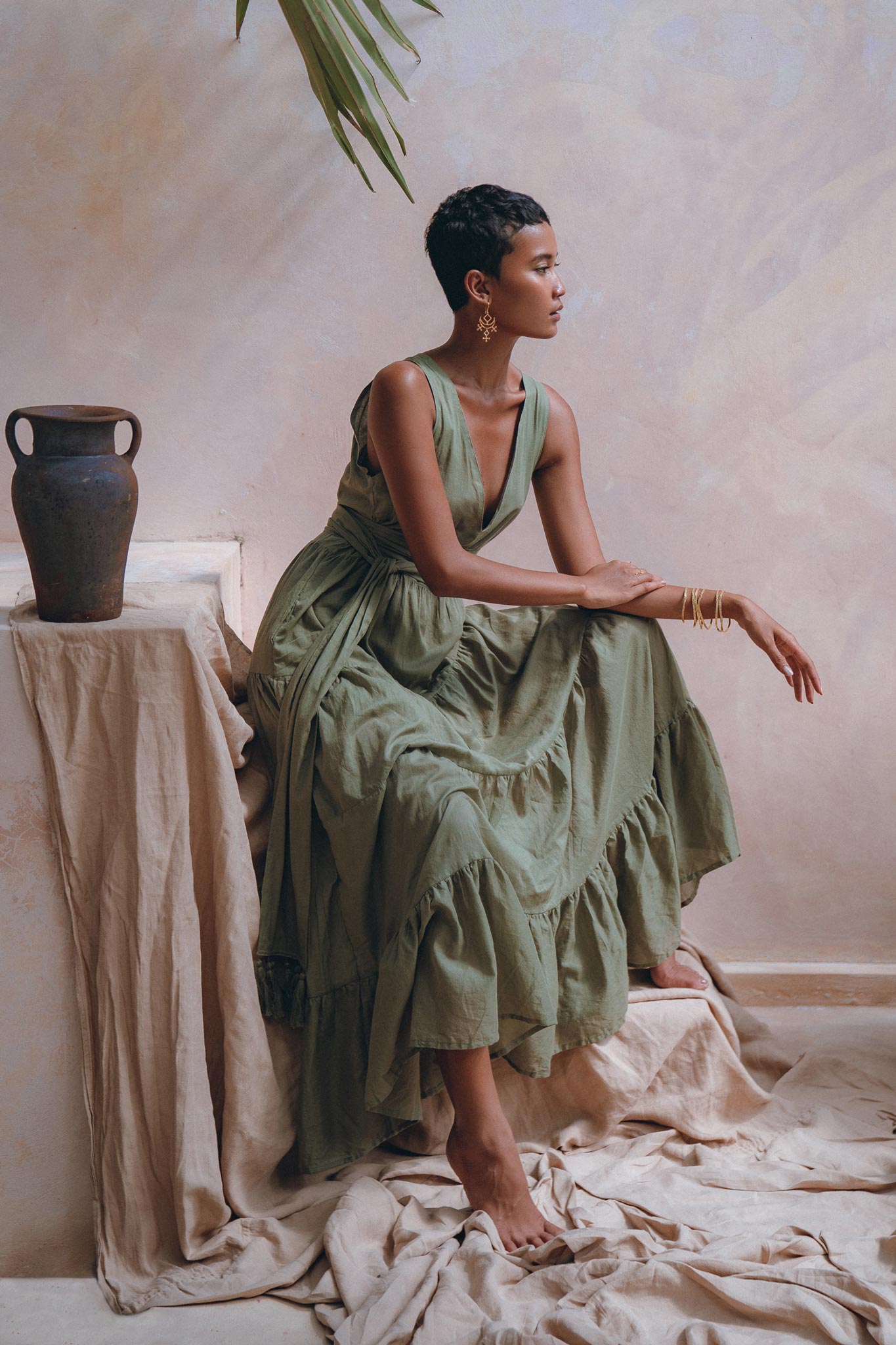 A woman in a flowing Sage Green Adjustable Dress from AYA Sacred Wear sits gracefully on a draped surface, next to a decorative vase. Her posture is elegant, and the soft lighting adds a serene ambiance. A large green leaf hangs above, enhancing the earthy setting.