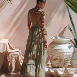 A person stands with their back to the camera, wearing a sage green Boho Bridesmaid Dress by AYA Sacred Wear. They are in a sunlit space adorned with large ceramic vases, beige fabric, and tropical leaves casting shadows around them.