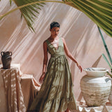 A person wearing the Boho Bridesmaid Dress by AYA Sacred Wear, featuring a bohemian style and adjustable sage green fabric, poses in an outdoor setting with large palm leaves casting shadows. Decorative vases and fabric on the ground enhance the artistic, tropical ambiance.