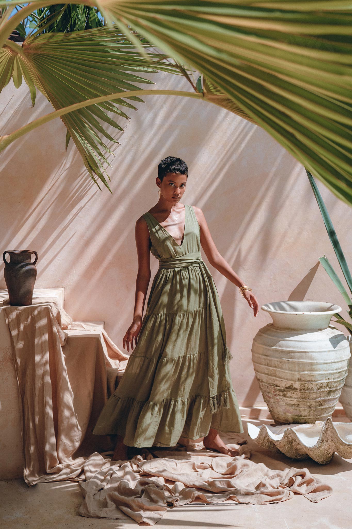 A person wearing the Boho Bridesmaid Dress by AYA Sacred Wear, featuring a bohemian style and adjustable sage green fabric, poses in an outdoor setting with large palm leaves casting shadows. Decorative vases and fabric on the ground enhance the artistic, tropical ambiance.