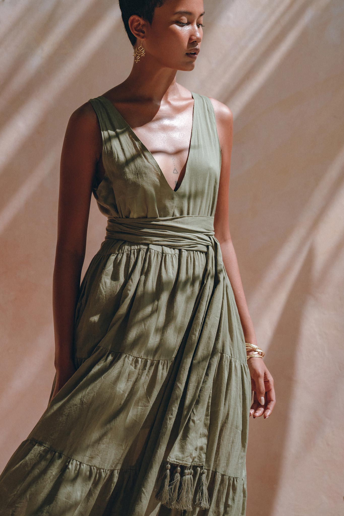 In soft lighting, a person is dressed in a sleeveless sage green Boho Bridesmaid Dress by AYA Sacred Wear. Crafted from organic cotton, the dress showcases a deep V-neck and tiered skirt, with an adjustable sash cinching the waist. The individual sports short hair adorned with earrings and bracelets, casting shadows on a light background.