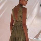 A person with short hair, adorned in the Boho Bridesmaid Dress by AYA Sacred Wear, elegantly stands in a sunlit spot accompanied by soft shadows on a light background. The sleeveless sage green adjustable dress highlights its open-back design and layered skirt as they look over their shoulder.