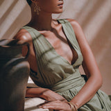 A woman in an AYA Sacred Wear Boho Bridesmaid Dress, a Sage Green Adjustable Bohemian Dress, sits beside a clay pot on a table. She has short hair and wears large earrings along with bracelets. Sunlight casts shadows across her and her surroundings, creating a dappled effect.